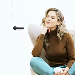 The picture shows a woman sitting in an armchair with her coffee cup. The door behind her is closed with the Griffwerk door handle Lucia smart2lock in graphite black.